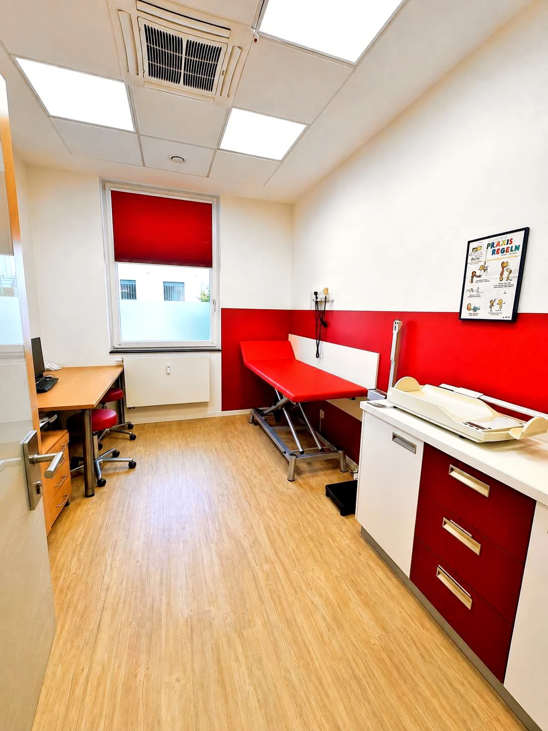 Behandlungszimmer mit roter Untersuchungsliege, Säuglingswaage und Arbeitsplatz